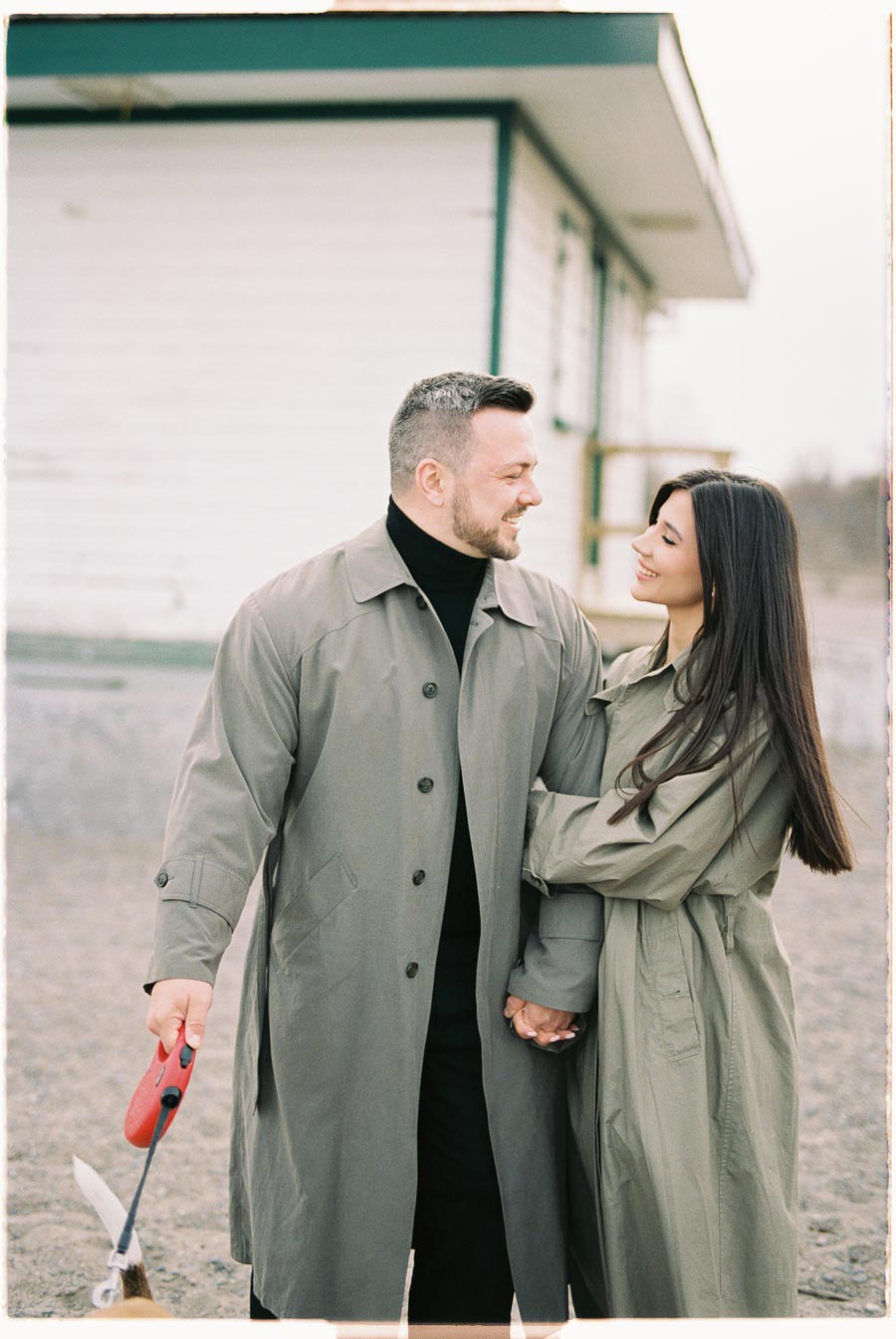 A couple in matching trench coats shares a moment of affection while holding hands outdoors.