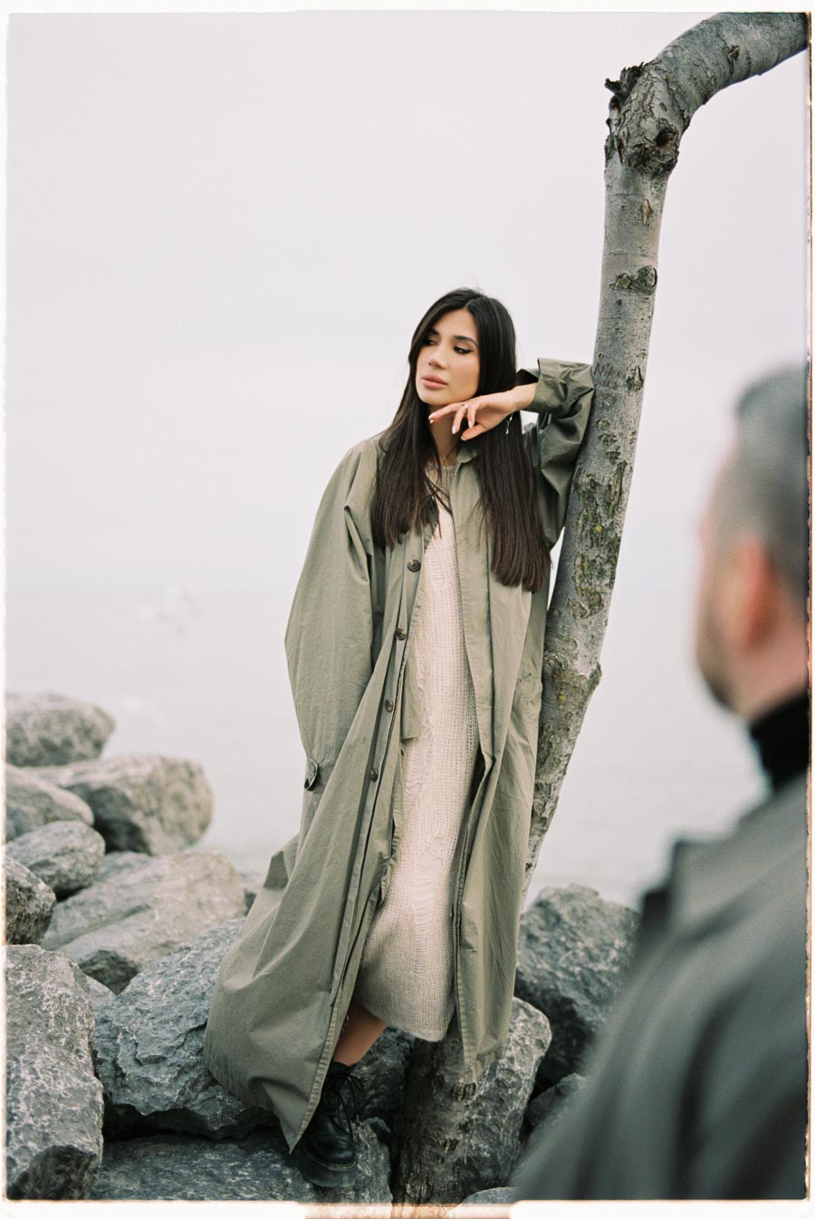 A woman in a long green coat leans against a tree by the sea, with rocks and a cloudy sky in the background.
