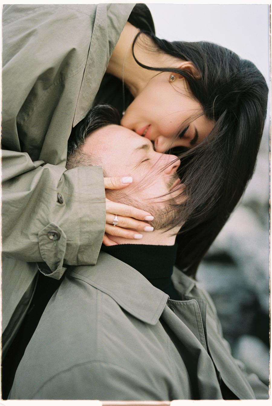 A tender moment between a couple, with the woman kissing the man's forehead while both wear matching green coats.