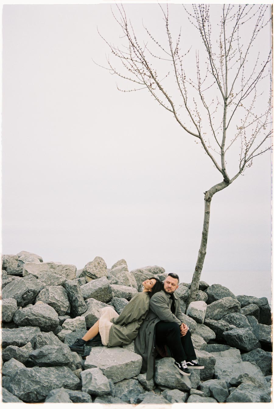 A serene moment captured as a couple sits on large rocks by a leafless tree, with the woman leaning on the man's back. The overcast sky adds a calm atmosphere to the scene.
