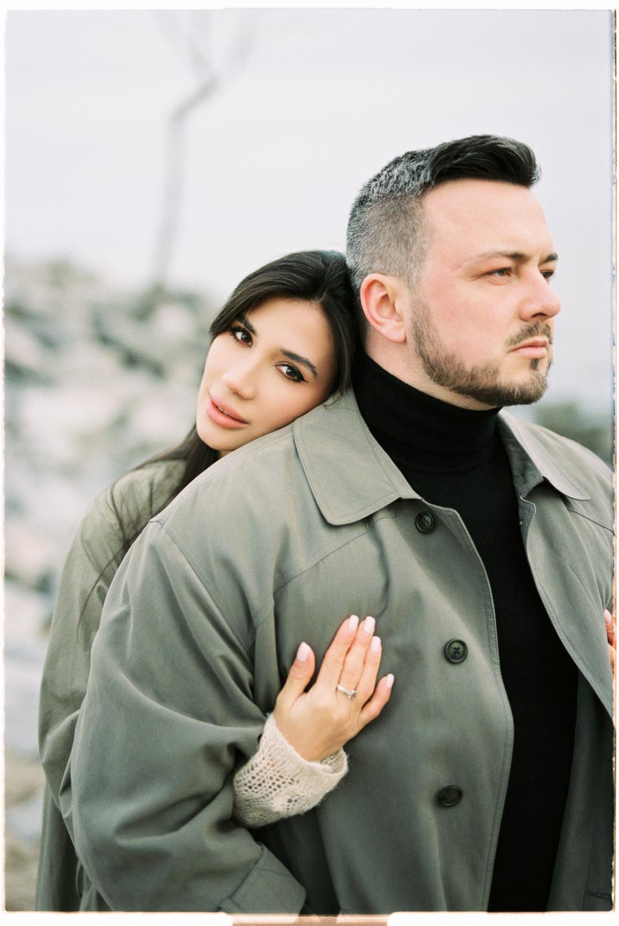 A couple embraces warmly in an outdoor setting, both dressed in stylish coats.