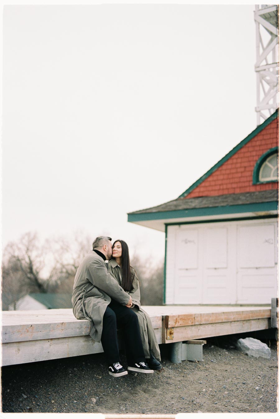 A couple shares a tender moment on a wooden platform, with a quaint building in the background.