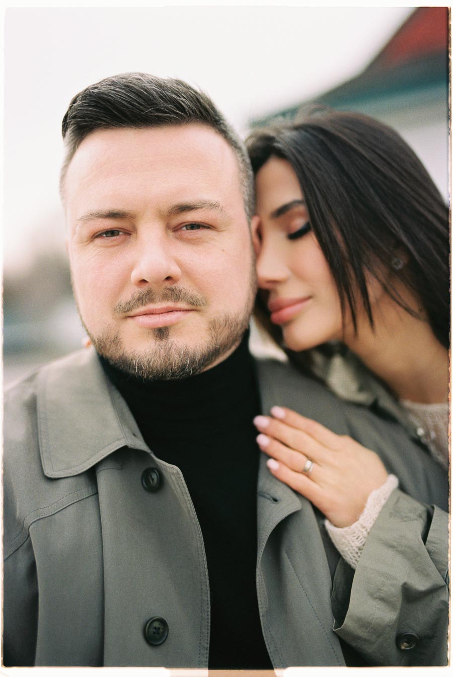 A close-up portrait of a couple, with the woman resting her head on the man's shoulder, showcasing an engagement ring.