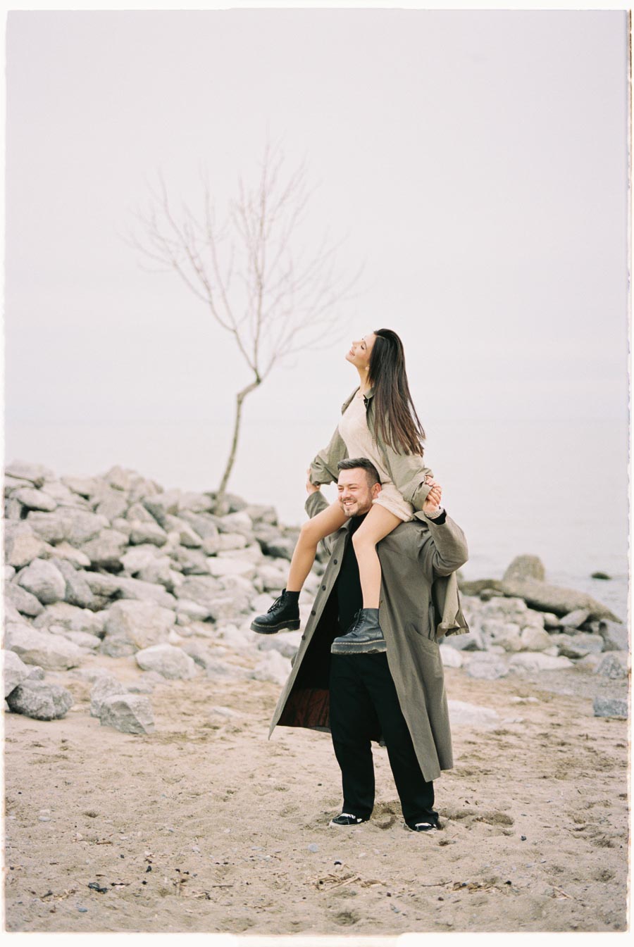 A joyful moment captured at a rocky beach, where a man carries a woman on his shoulders. Both are dressed in long coats, with the woman looking up blissfully. A bare tree and calm water are visible in the background, adding to the serene atmosphere.