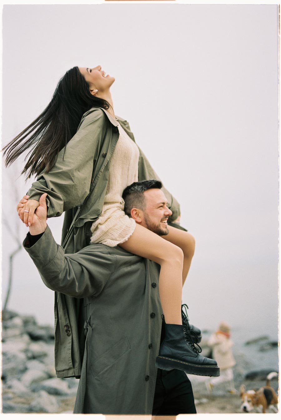 A joyful moment captured as a woman sits on a man's shoulders, both dressed in green coats, enjoying a day at the beach.