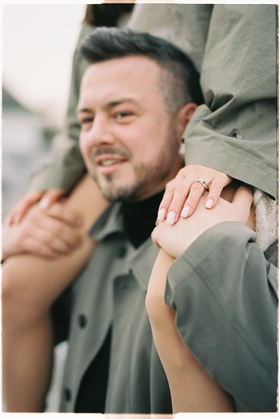 A man smiling while carrying someone on his shoulders, with the person's hands resting on his head, displaying a ring.
