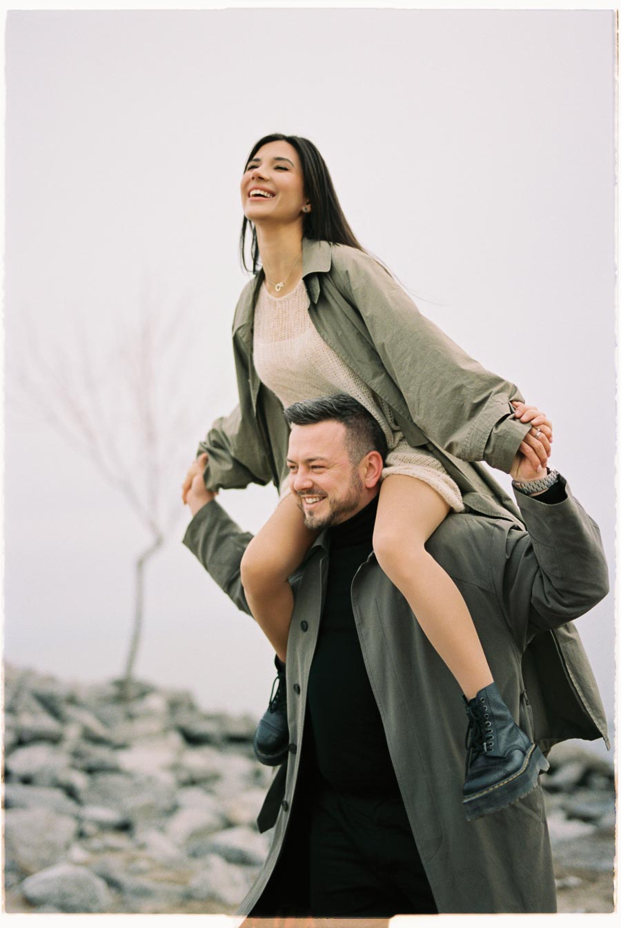 A joyful moment captured as a woman sits on a man's shoulders, both smiling broadly. They are dressed in casual coats, with a rocky landscape in the background, suggesting a chilly day outdoors.