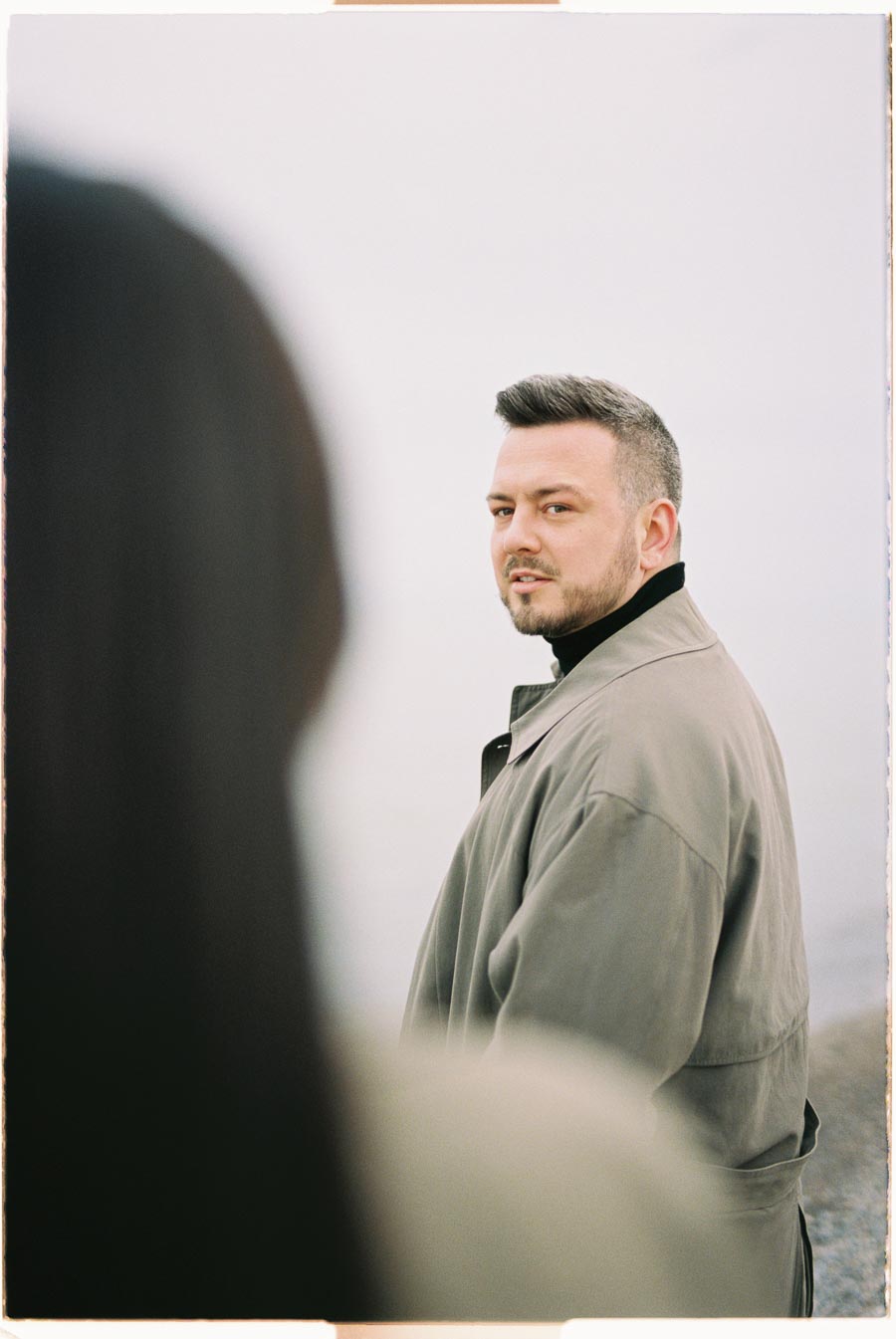 A man in a coat looks over his shoulder, with a blurred figure in the foreground, creating a sense of depth.