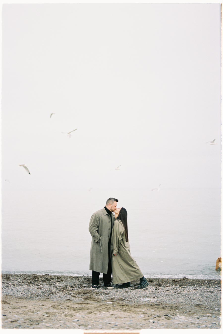 A couple shares a kiss on a serene beach, both wearing long coats, with seagulls flying overhead.