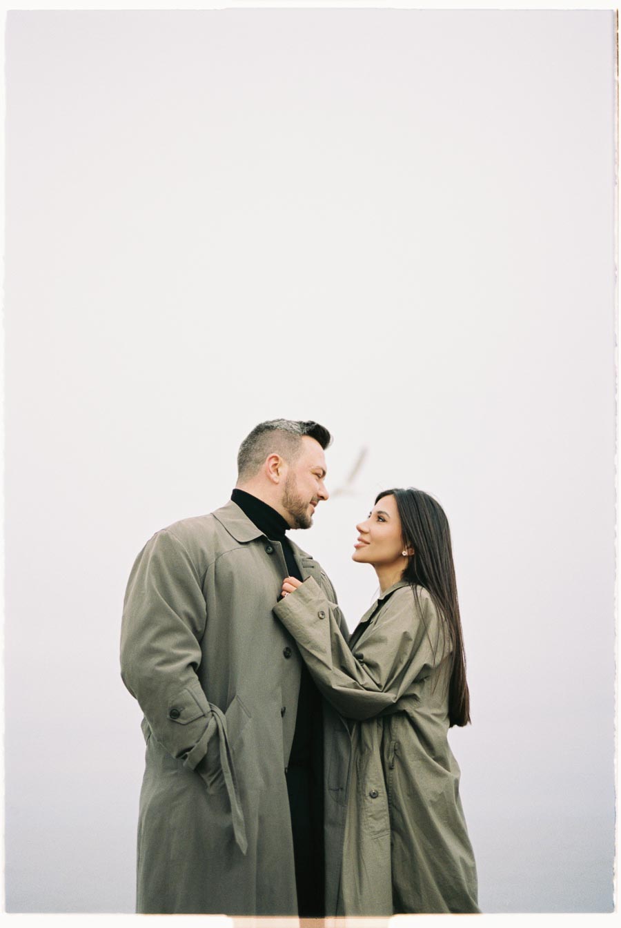 A couple in matching trench coats share a tender moment, gazing into each other's eyes against a minimalist backdrop.