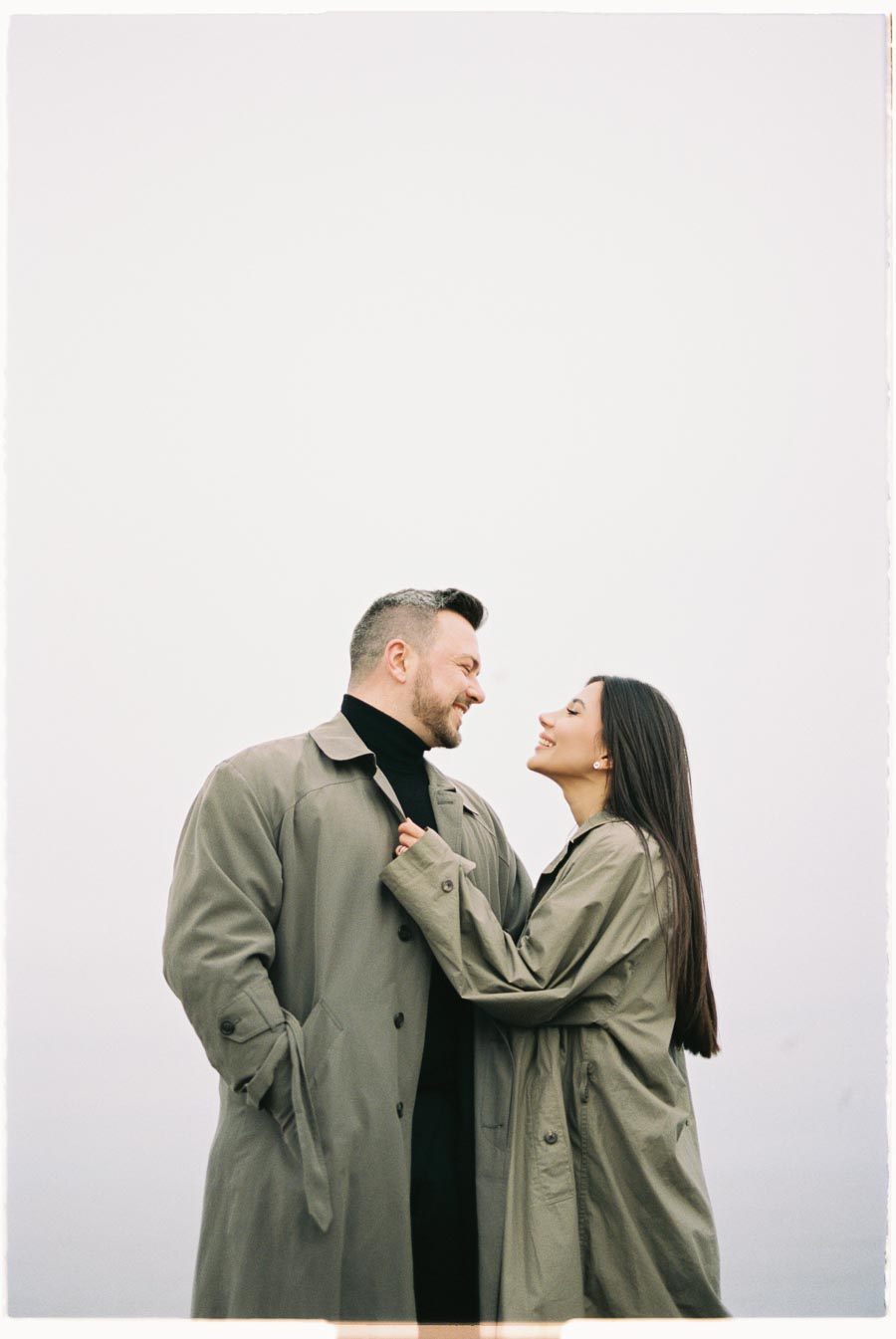 A couple in matching trench coats share a joyful moment, smiling at each other against a minimalist backdrop.