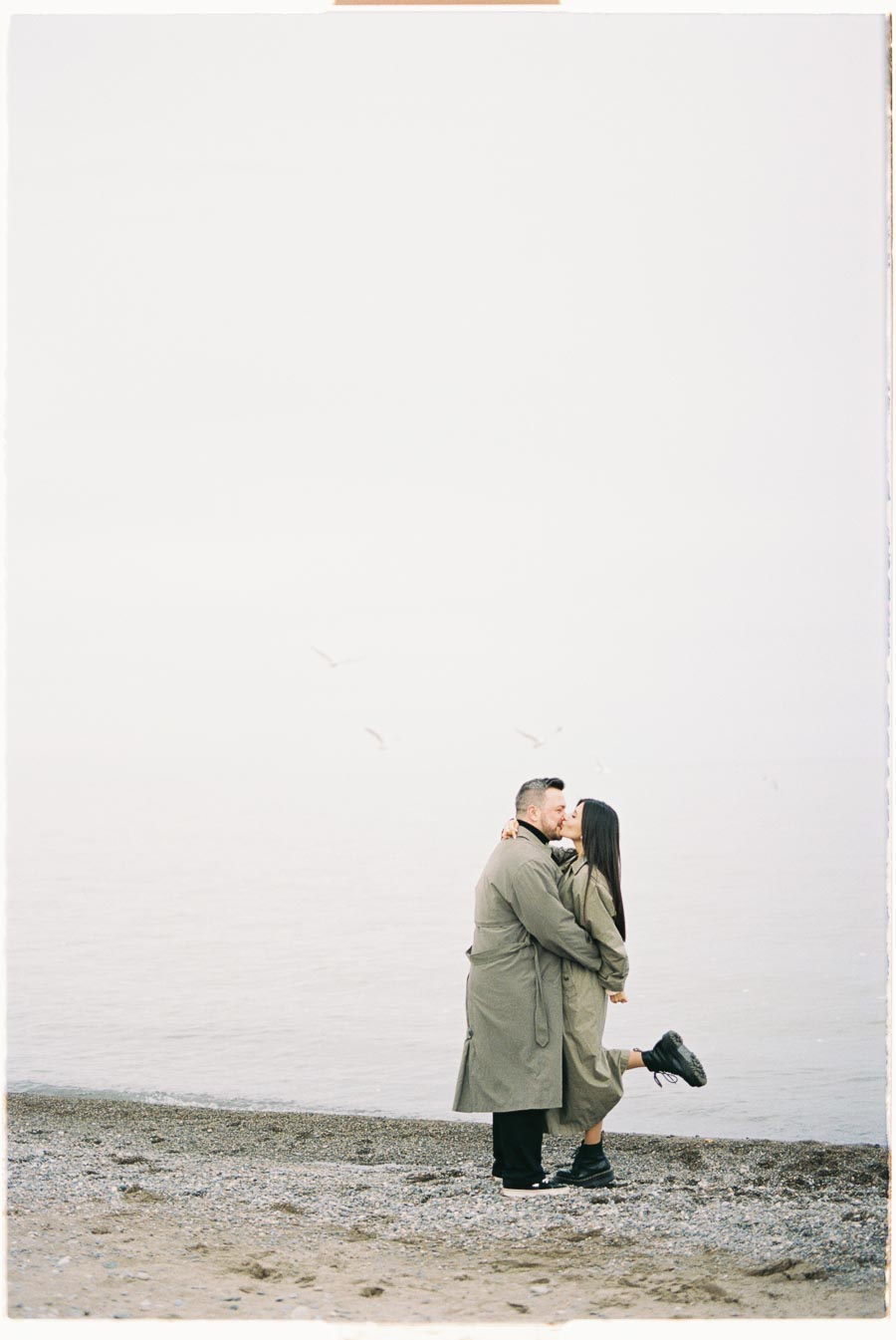 A couple embraces on a foggy beach, both wearing matching trench coats. The serene scene is complemented by seagulls flying in the background.