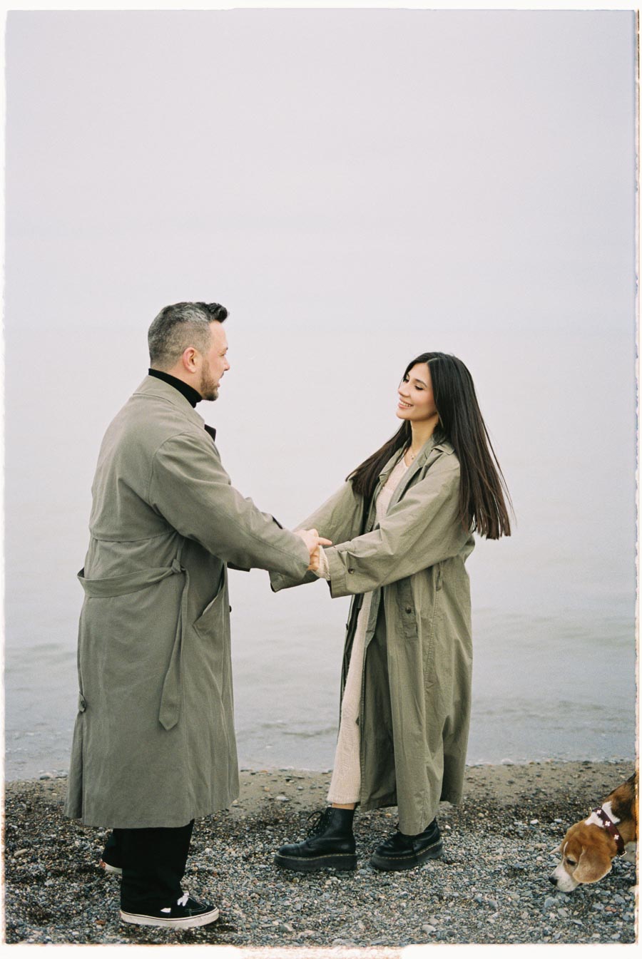 A couple in matching trench coats hold hands by the water, accompanied by a beagle.
