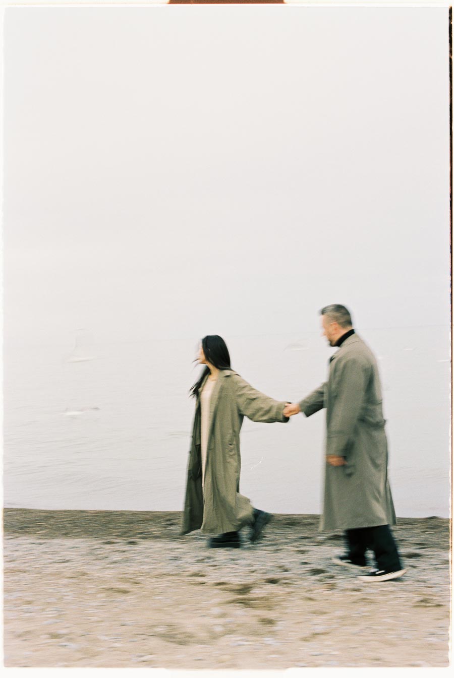 A couple in long coats walks hand in hand along a beach, captured in a blurred motion.