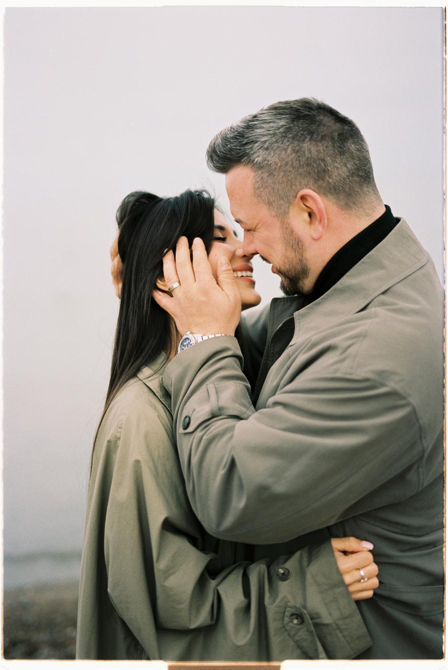 A couple shares a tender moment, embracing and smiling at each other while wearing matching beige coats.