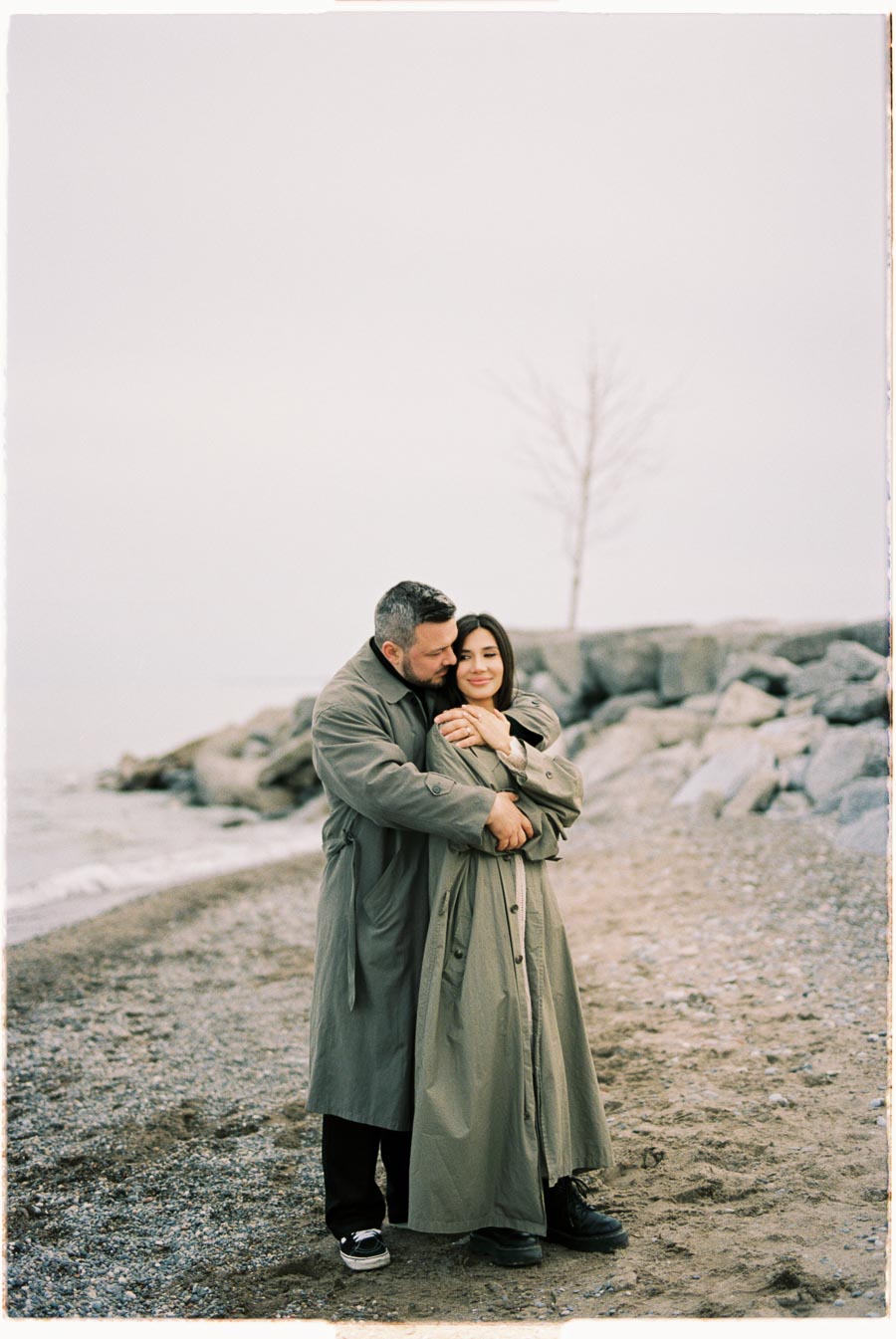 A couple embraces on a beach, both wearing matching trench coats, with a rocky shoreline and a bare tree in the background.