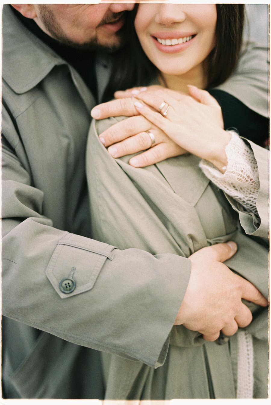 A close-up of a couple embracing, showcasing their hands and smiles. Both are dressed in light-colored coats, with the woman wearing a ring.