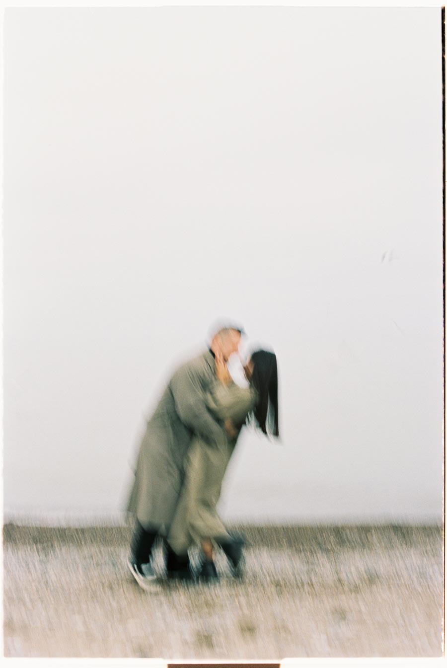 A blurred photograph captures two people in an embrace, sharing a kiss while wearing long coats. The background is plain, emphasizing the motion and emotion of the moment.