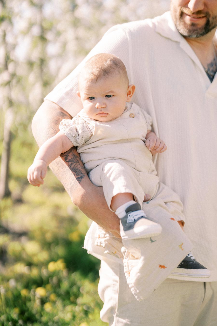 A baby in light clothing and sneakers is held by an adult in a sunny outdoor setting.