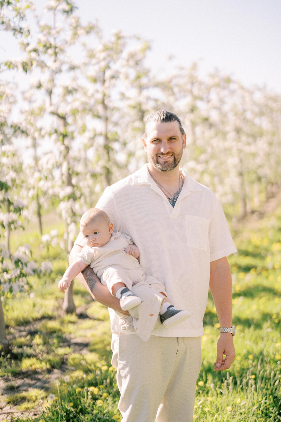 A man smiles while holding a baby in a sunny orchard with blooming trees.