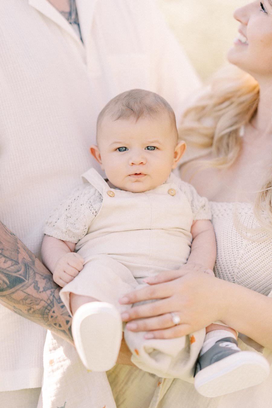 A baby in light-colored overalls is held by two adults, one with a tattooed arm and the other with long blonde hair, creating a warm family moment.