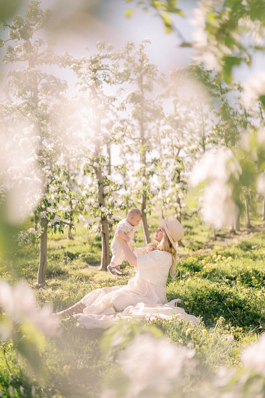 A serene moment in a blooming orchard as a woman in a white dress and hat joyfully holds a baby, surrounded by lush greenery and blossoms.
