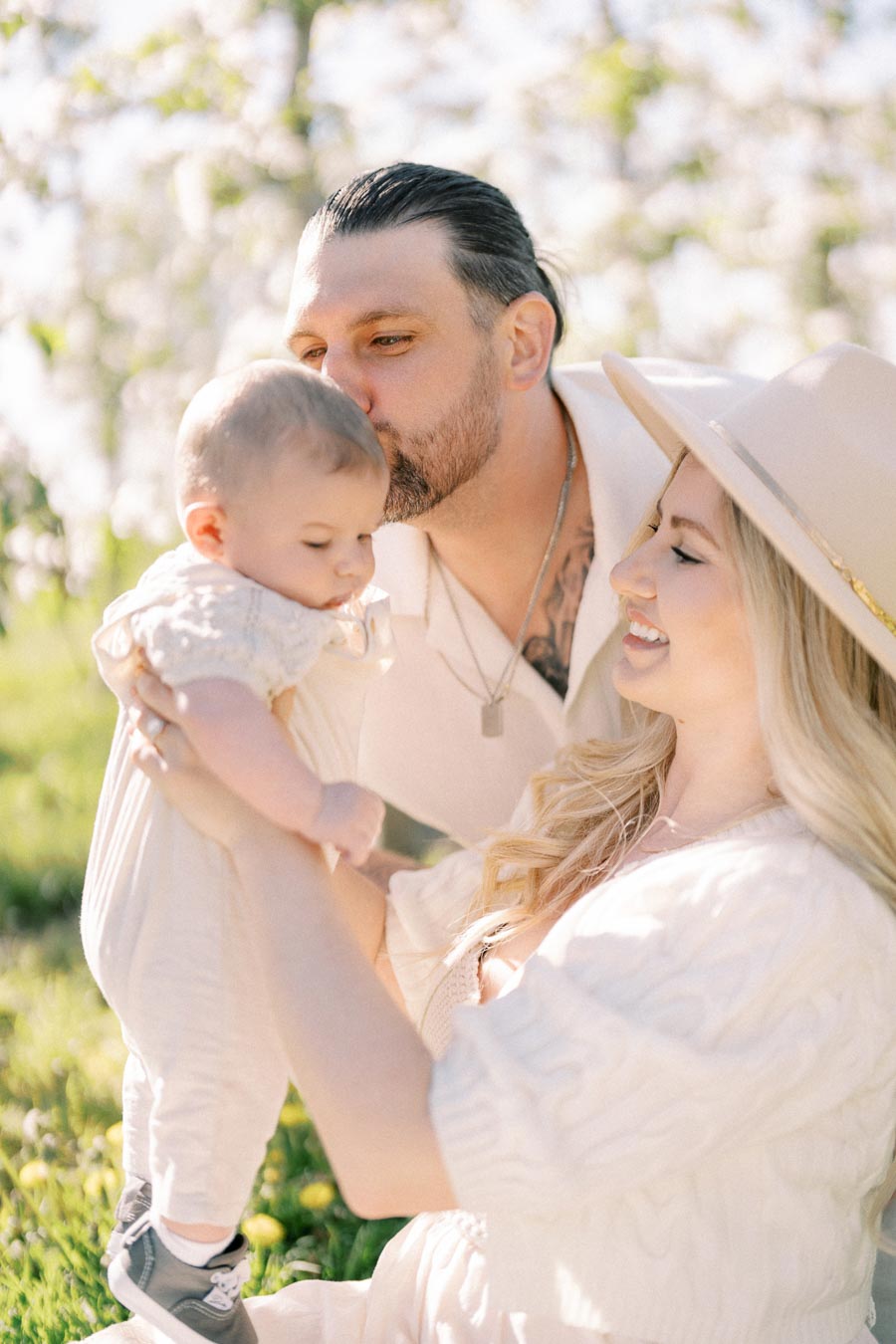 A family enjoying a sunny day outdoors, with a man kissing a baby held by a woman wearing a hat.