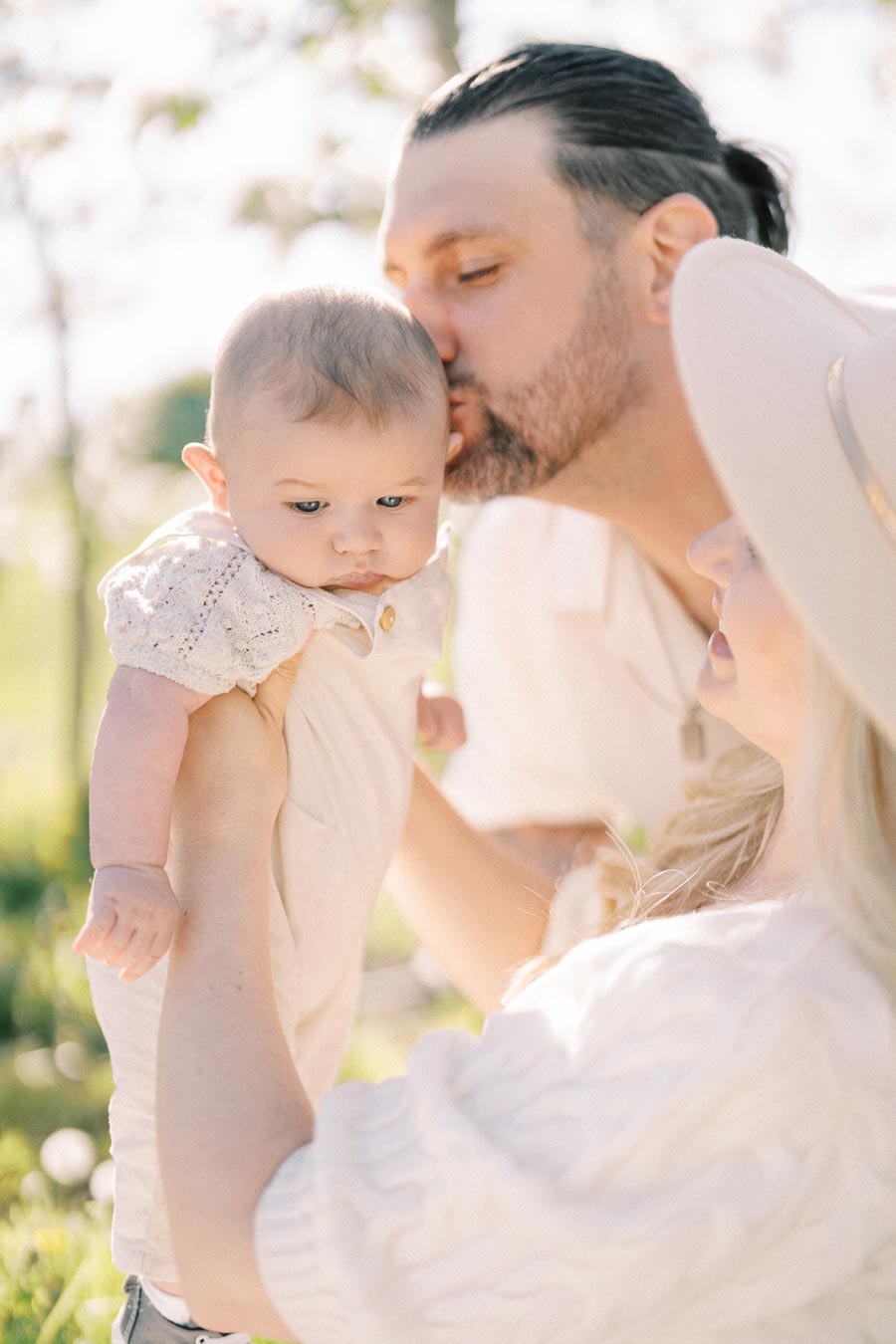 A tender family moment captured outdoors, with a baby being lovingly held by a woman as a man kisses the baby's head. The soft sunlight and greenery create a serene atmosphere.
