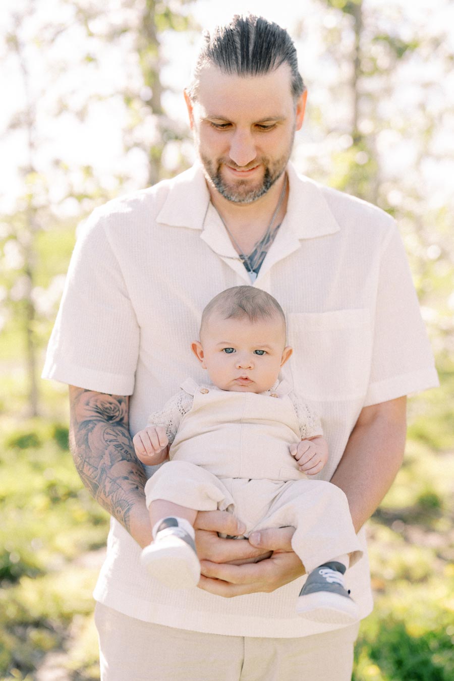 A man with tattoos holds a baby in a serene outdoor setting, both dressed in light-colored outfits.