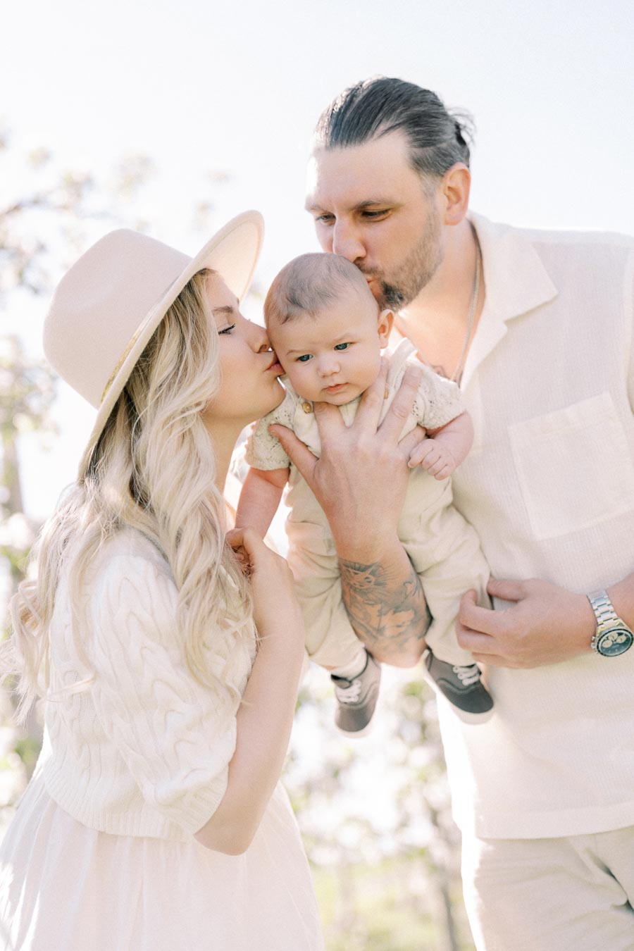 A couple lovingly holds and kisses their baby in a serene outdoor setting.