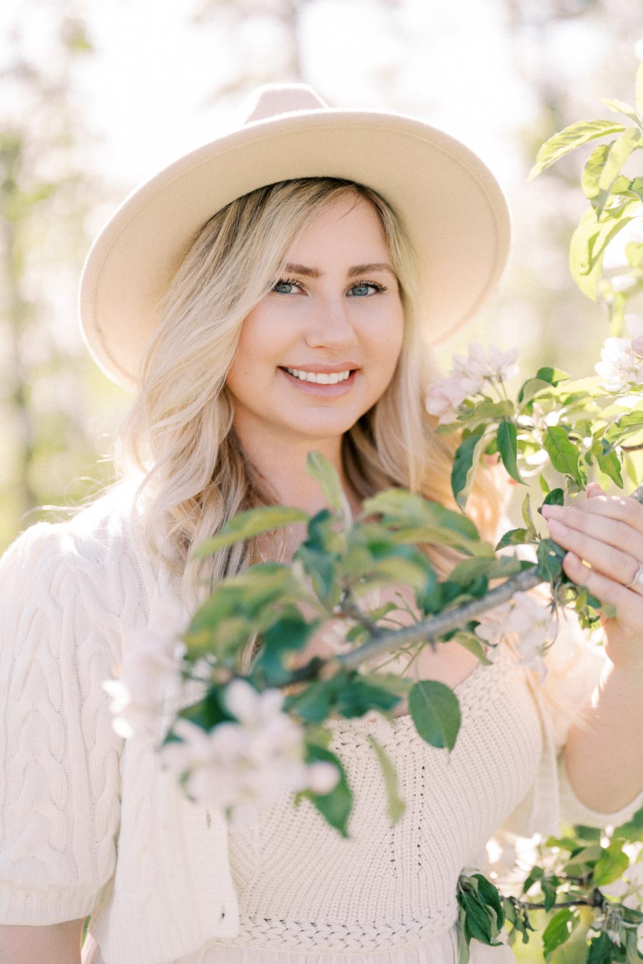 A woman in a white hat and sweater stands near a flowering tree branch, smiling in a sunlit outdoor setting.