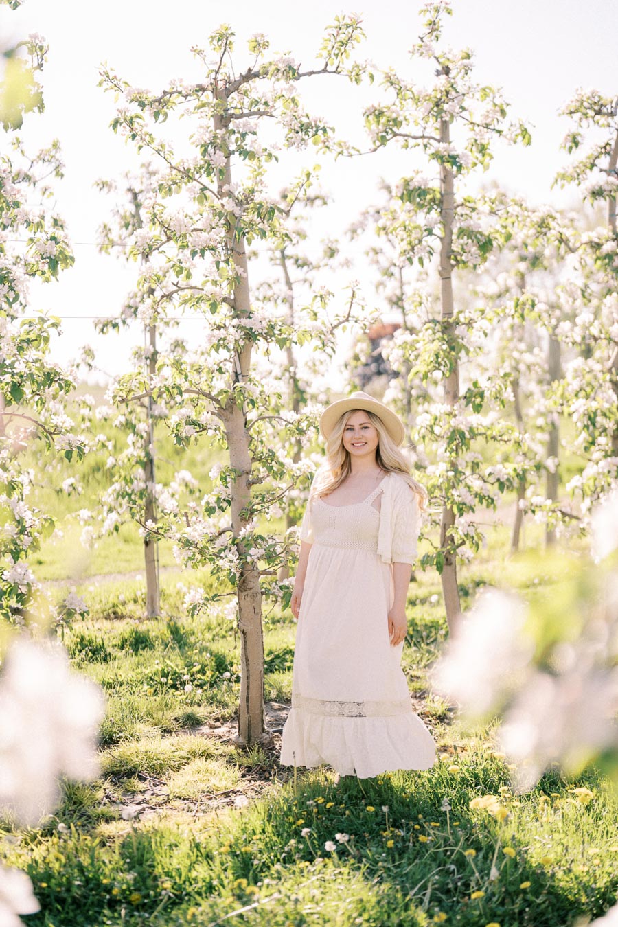A woman in a white dress and hat stands amidst blooming trees in an orchard, enjoying a sunny day.