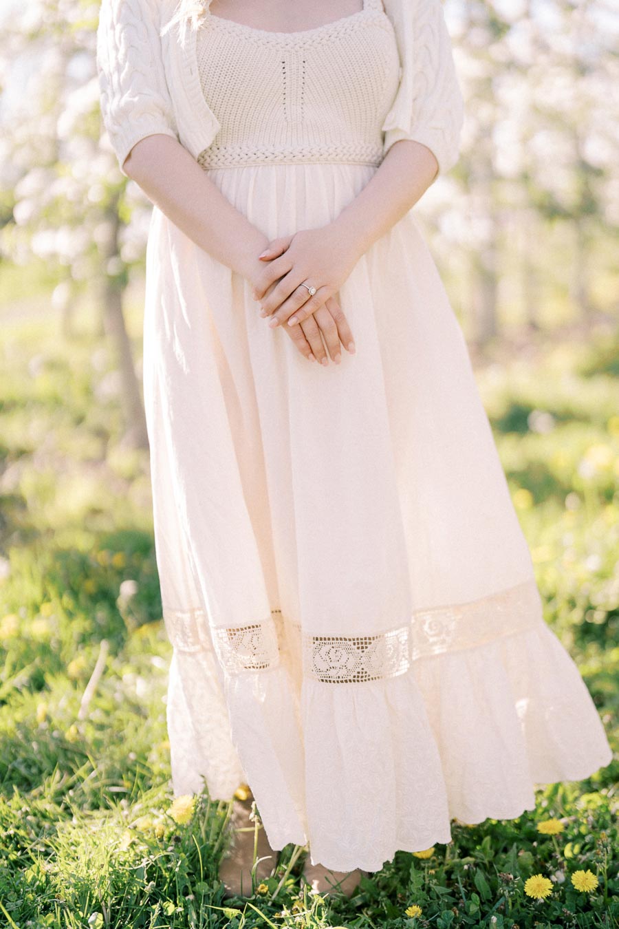 A person stands in a sunlit field wearing a light-colored dress and cardigan, with hands gently clasped.