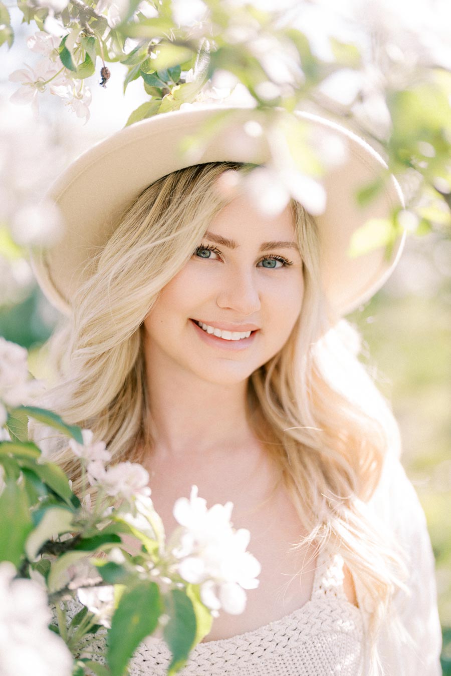 A woman with long blonde hair and a beige hat smiles among blooming branches, capturing a serene and sunny moment.