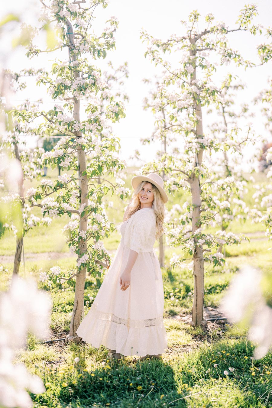 A woman in a white dress and hat stands gracefully among blooming trees in an orchard, enjoying a sunny day.