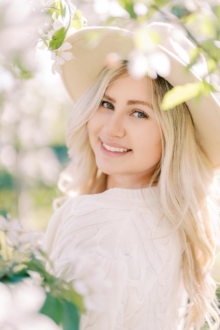 A woman smiles warmly while surrounded by blooming flowers, wearing a white sweater and a wide-brimmed hat.