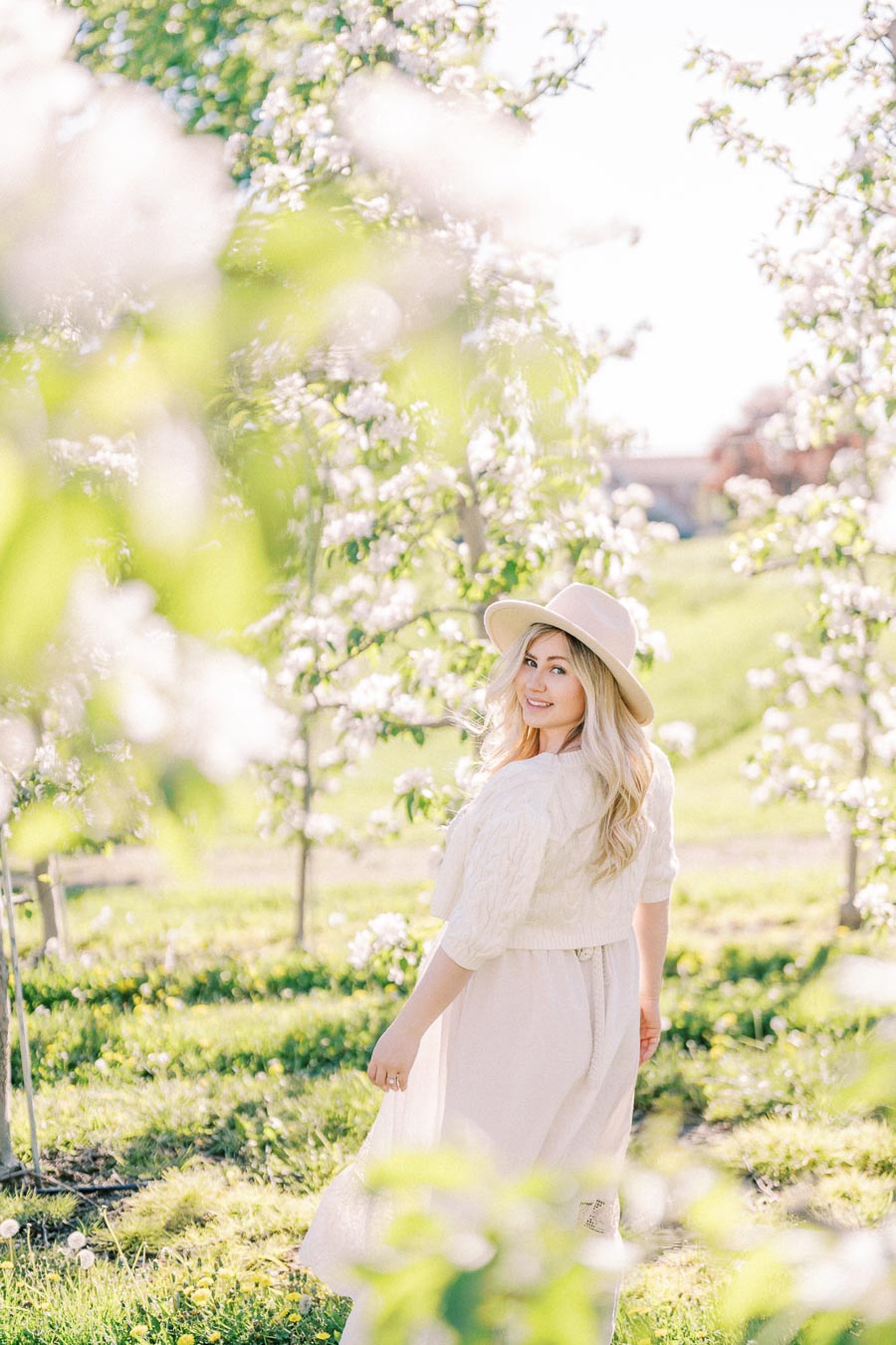 A woman in a white dress and hat stands in a blooming orchard, surrounded by lush greenery and white flowers, enjoying a sunny day.