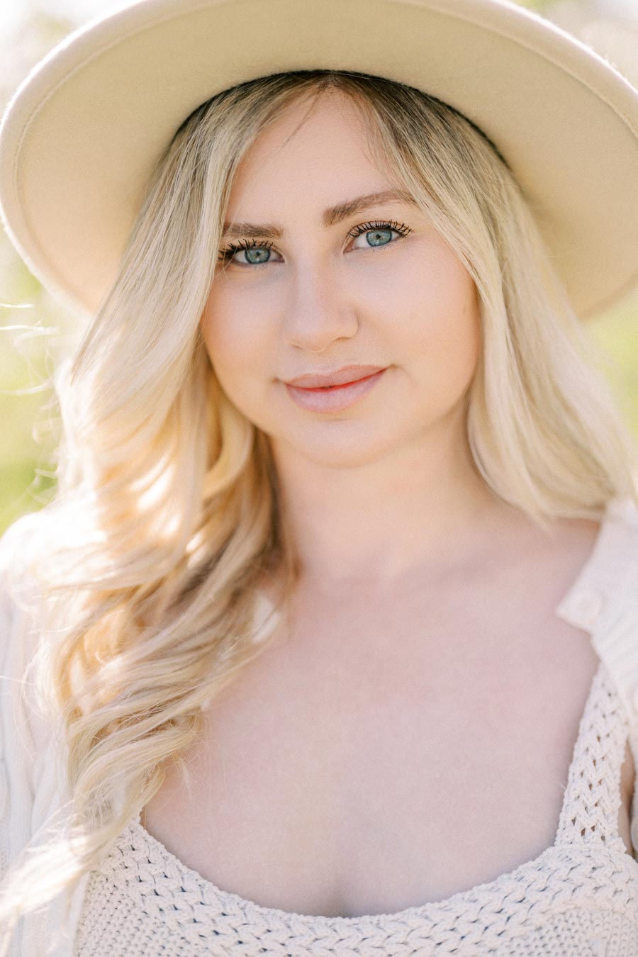 A woman with long blonde hair wearing a beige hat and a knitted top, smiling softly outdoors.