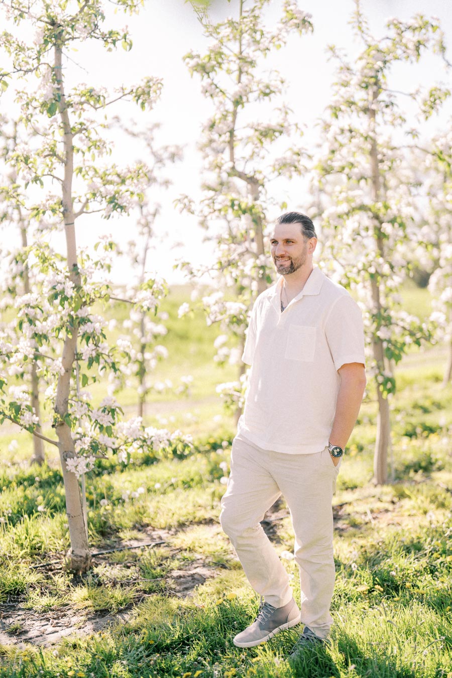 A man enjoys a sunny day in an orchard filled with blooming trees, dressed in light-colored casual attire.