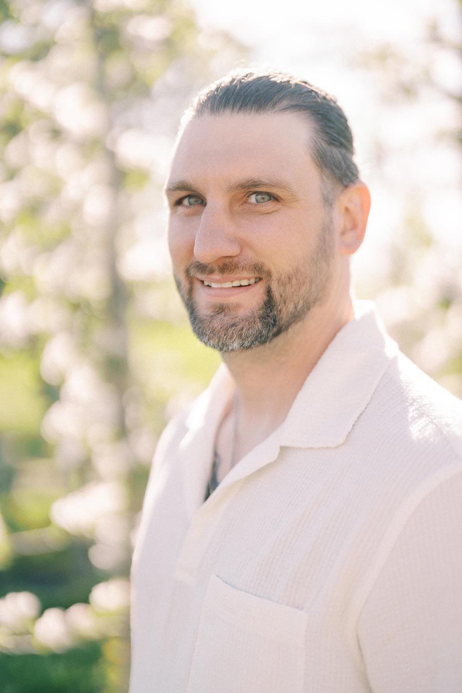 A man smiling outdoors with a blurred background of greenery and flowers.
