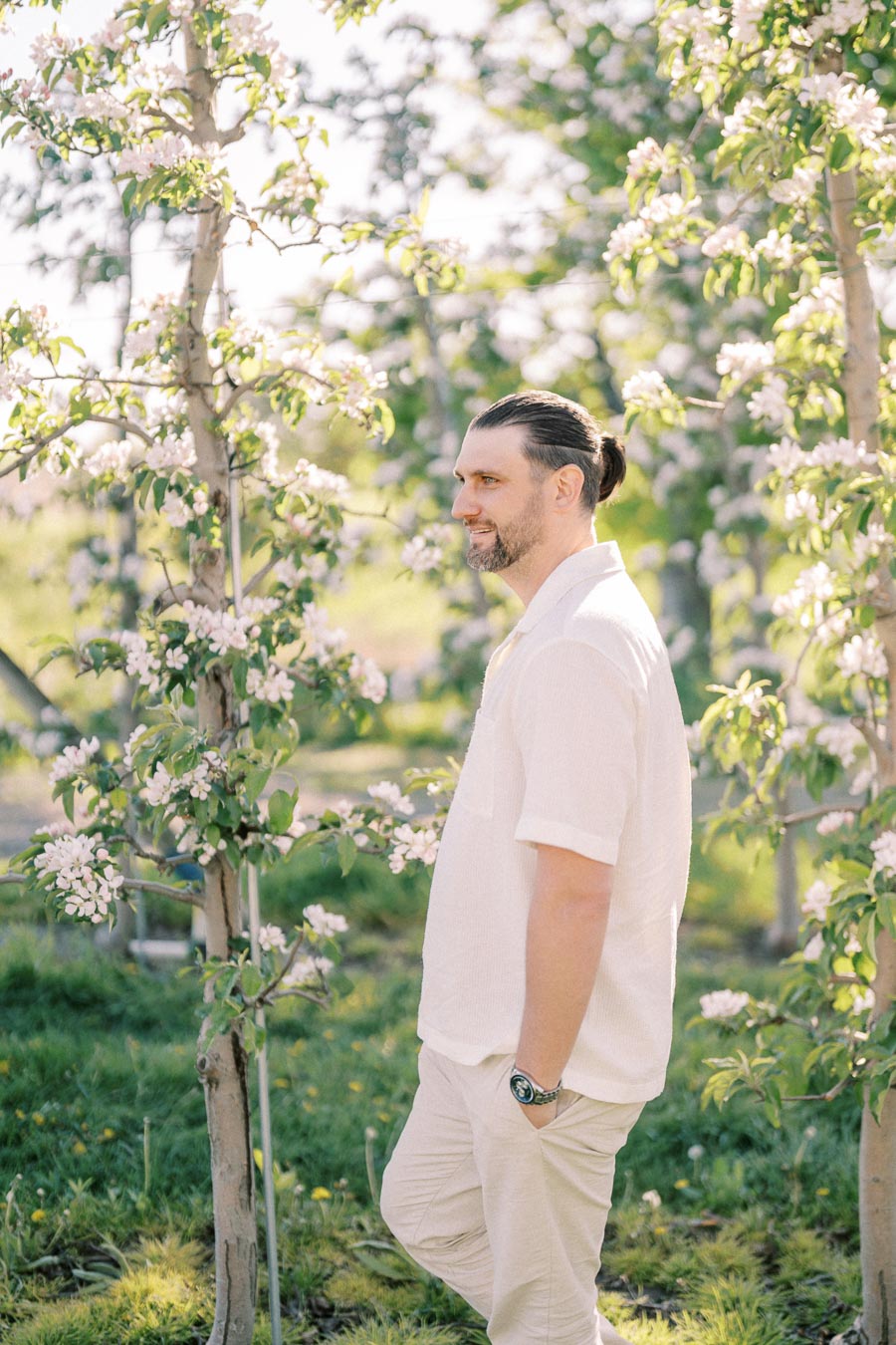 A man in a white shirt stands in an orchard surrounded by blooming trees, enjoying a sunny day.
