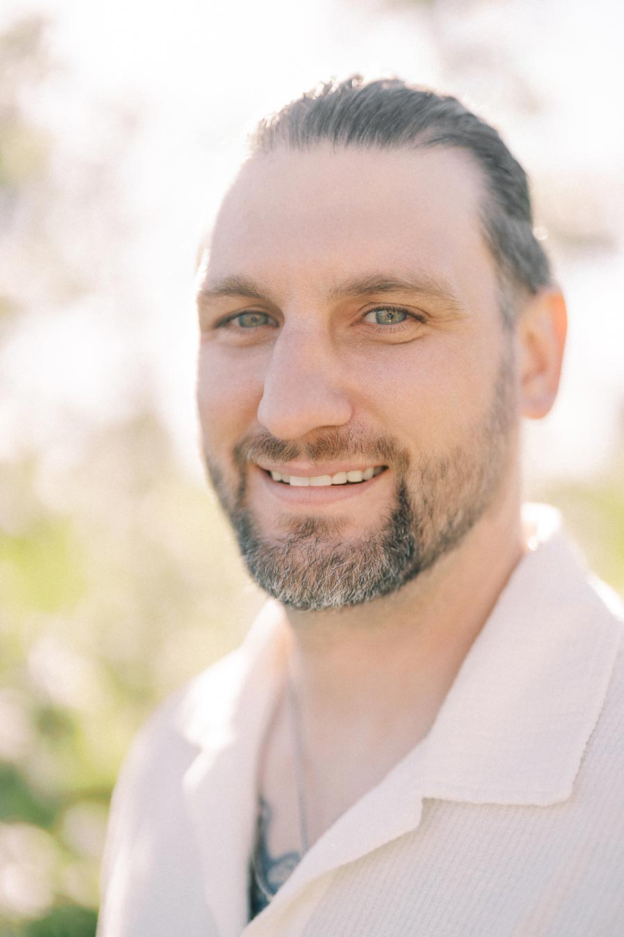 A man with a beard smiling outdoors in soft sunlight, wearing a light-colored shirt.
