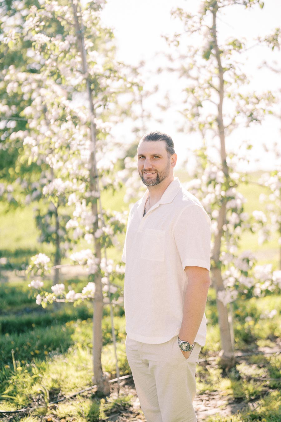 A man wearing a white shirt stands outdoors beside blooming trees, enjoying a sunny day.