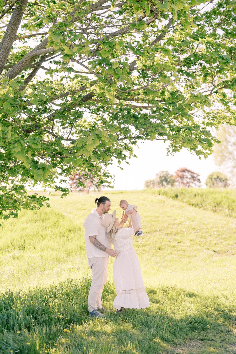 A joyful family moment captured under the shade of a tree in a sunny park, with the mother lifting the baby and the father standing close.