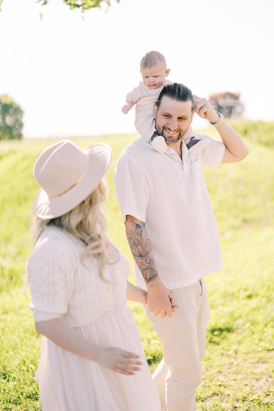 A family enjoying a sunny day outdoors, with a man carrying a baby on his shoulders and holding hands with a woman wearing a hat.