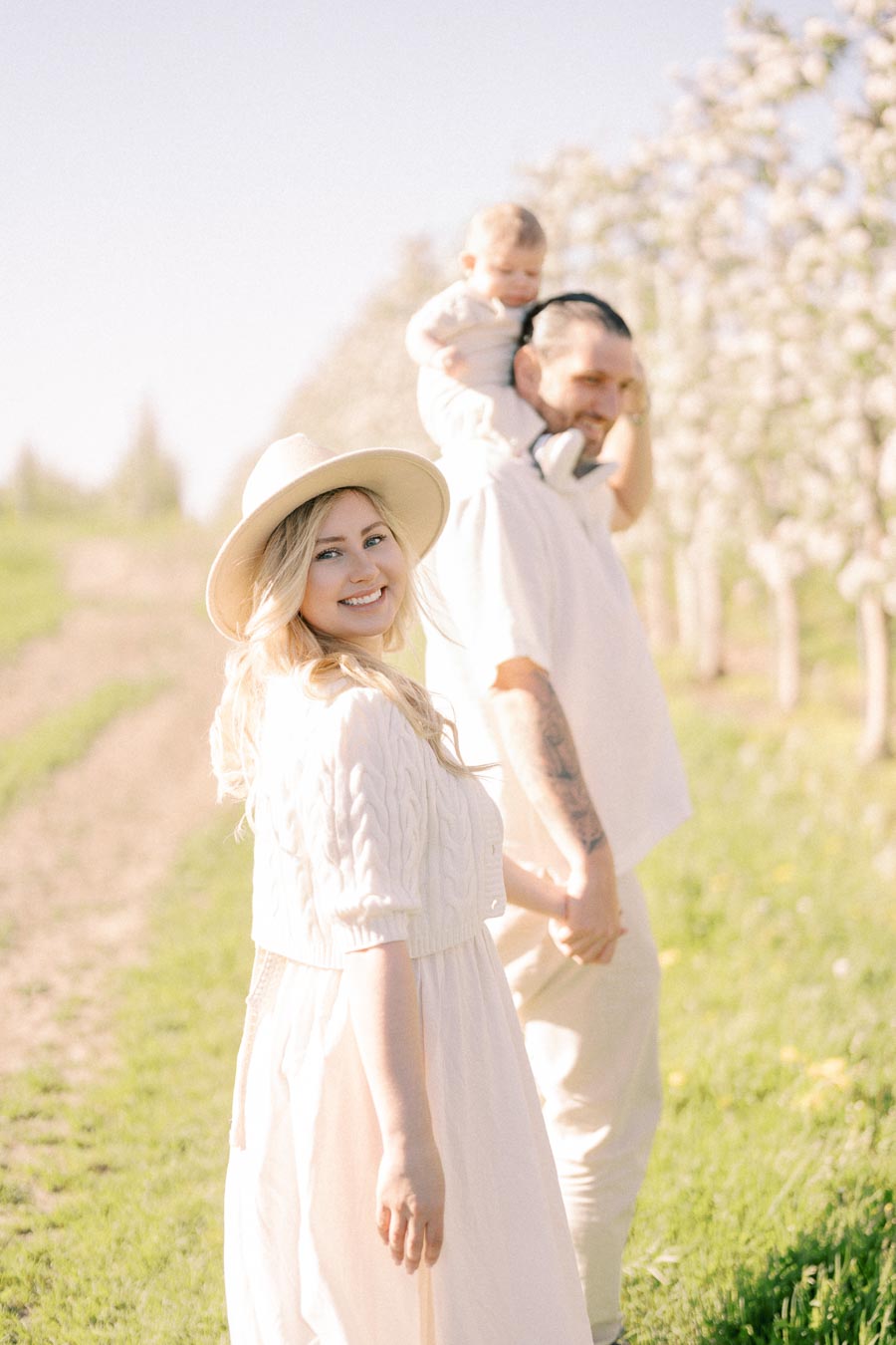 A family enjoys a sunny day in a blooming orchard, with the woman smiling at the camera while the man carries a child on his shoulders.