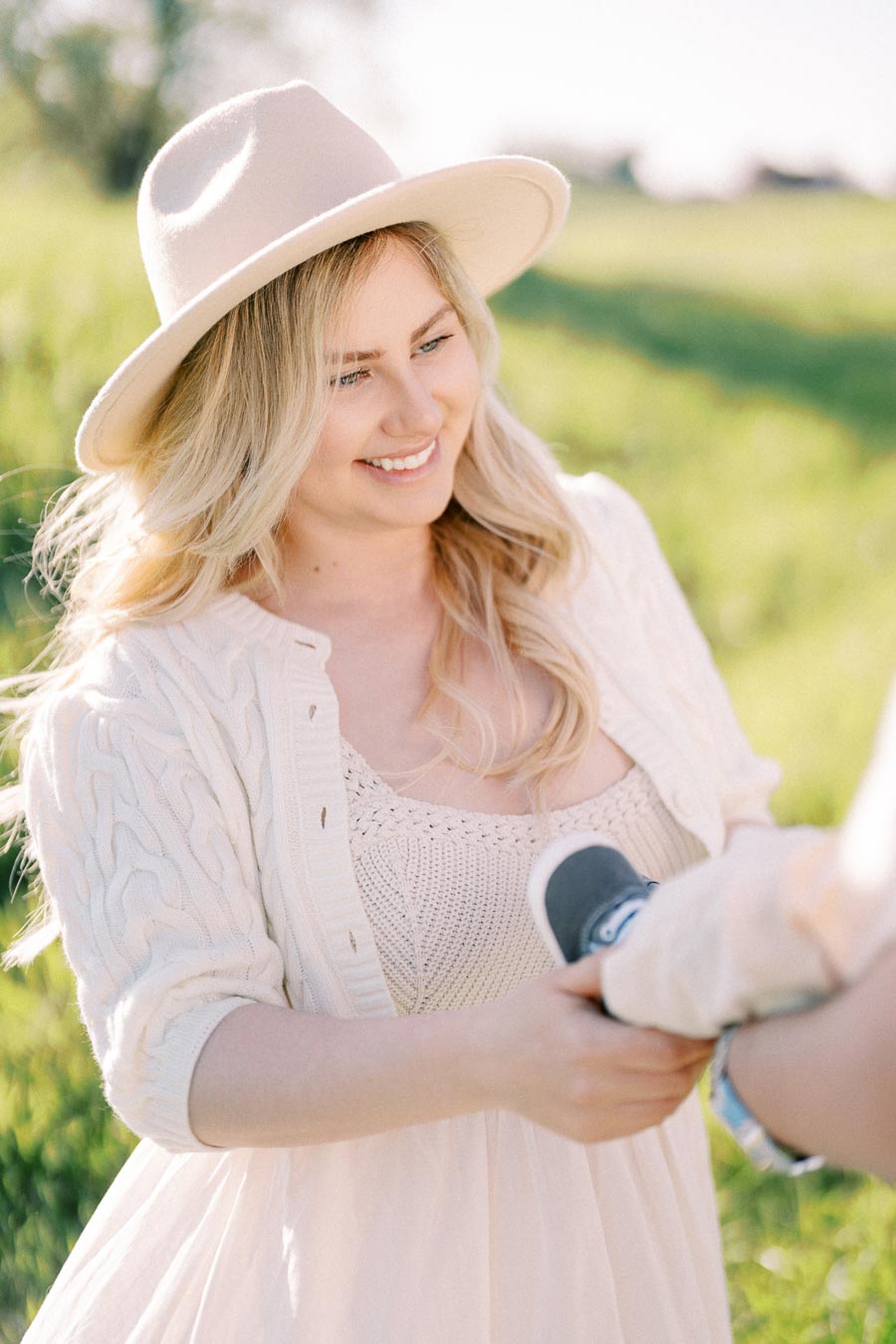 A woman in a white hat and sweater smiles while holding a baby's foot in a sunny, grassy field.