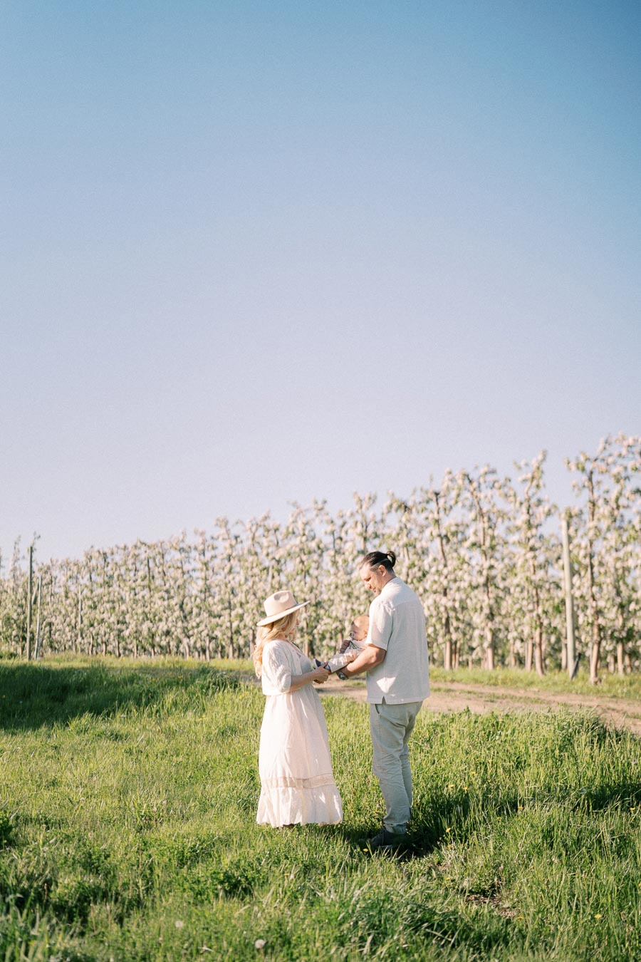 A couple stands in a grassy field holding a baby, surrounded by blooming trees under a clear blue sky.