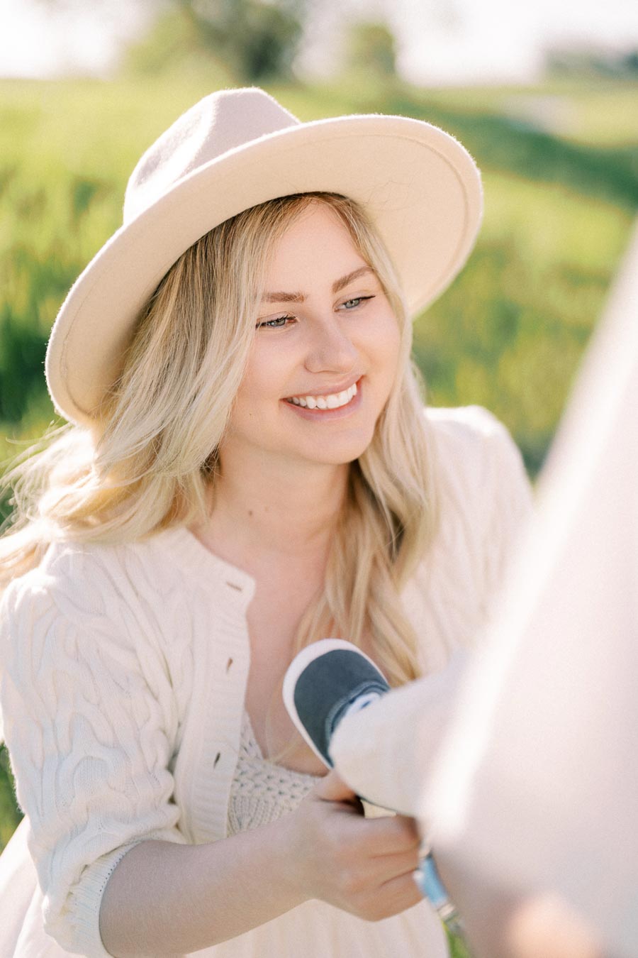 A woman in a light-colored hat and sweater smiles warmly while outdoors in a grassy field.