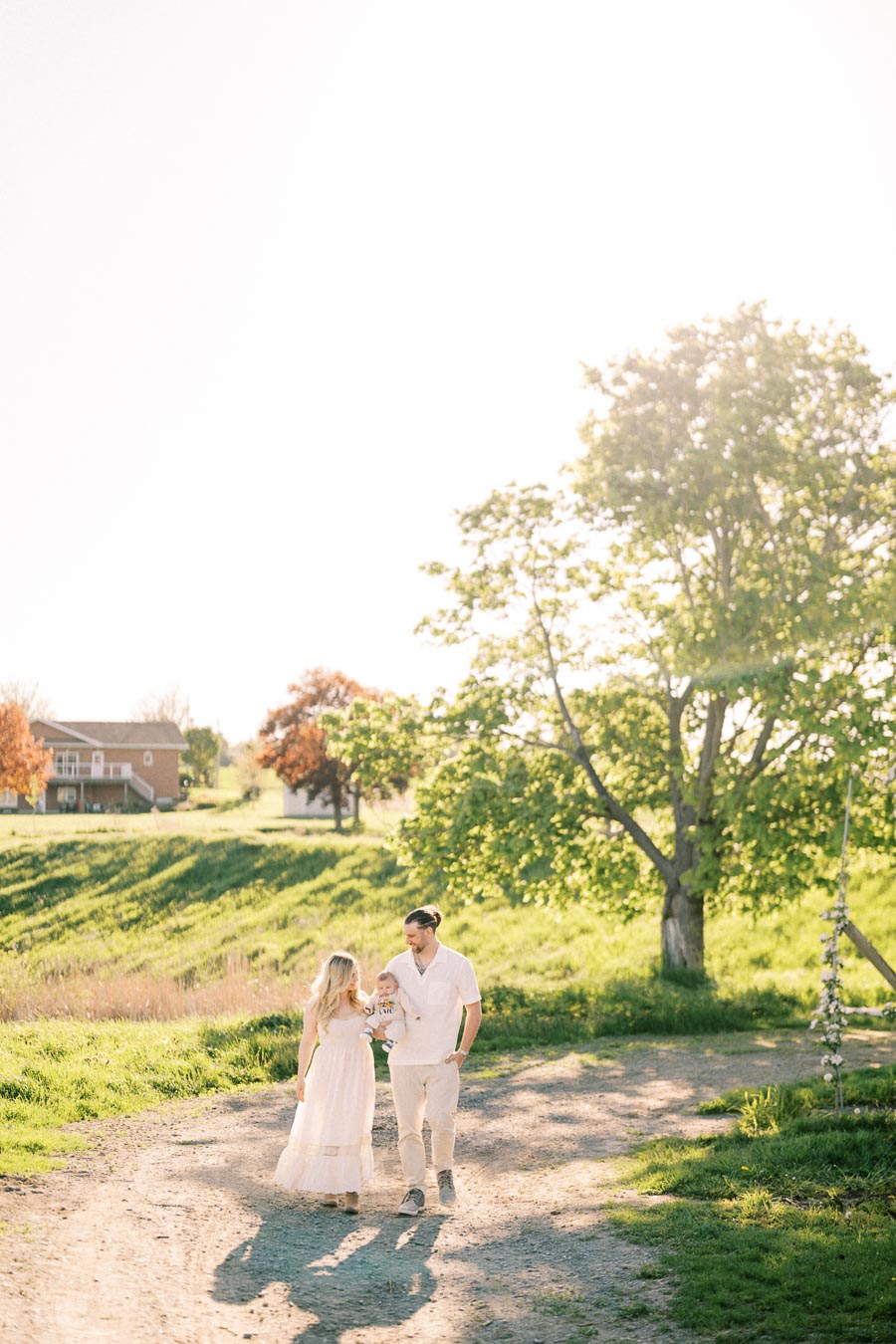A couple enjoys a sunny walk with their baby in a picturesque rural setting, surrounded by lush greenery and a distant house.